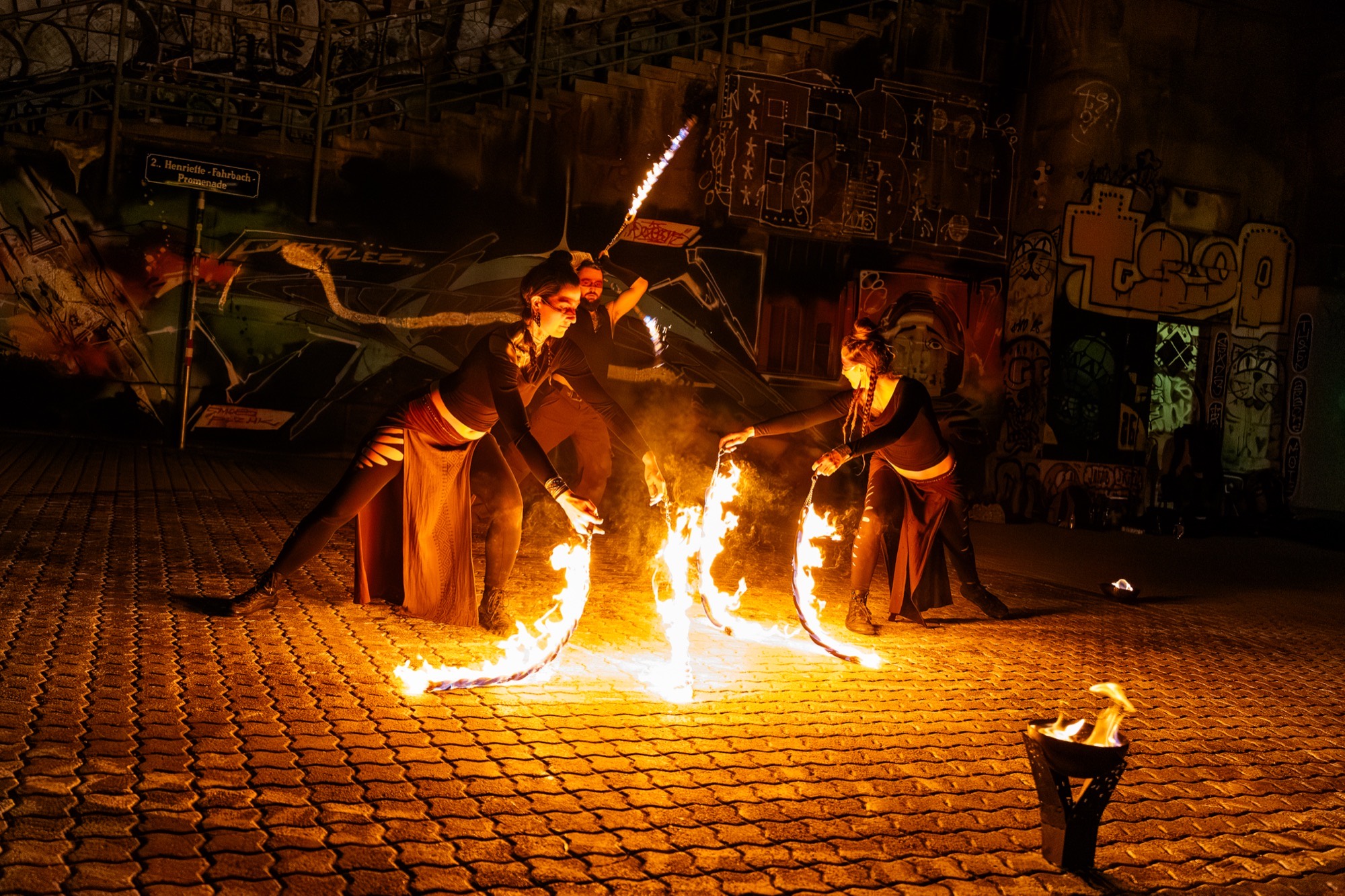 Three fire performers spinning fire ropes and staff before a graffiti wall