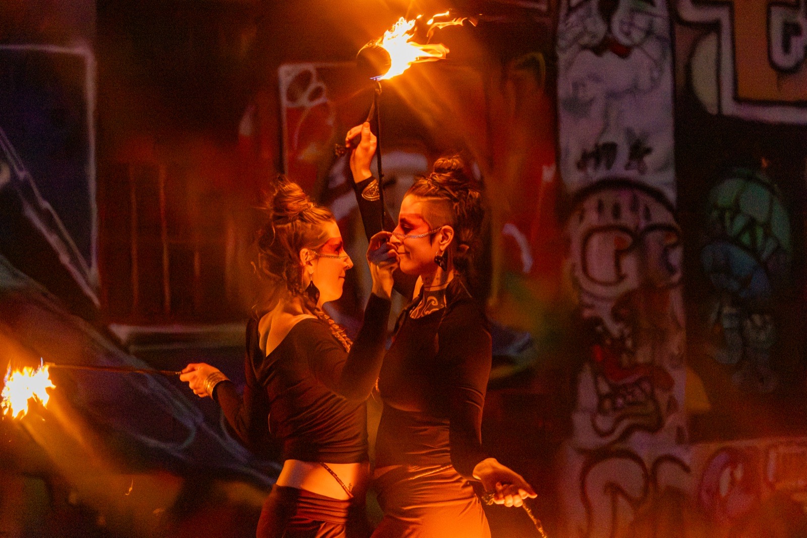Two fire performers with a burning prop by a graffiti wall at night